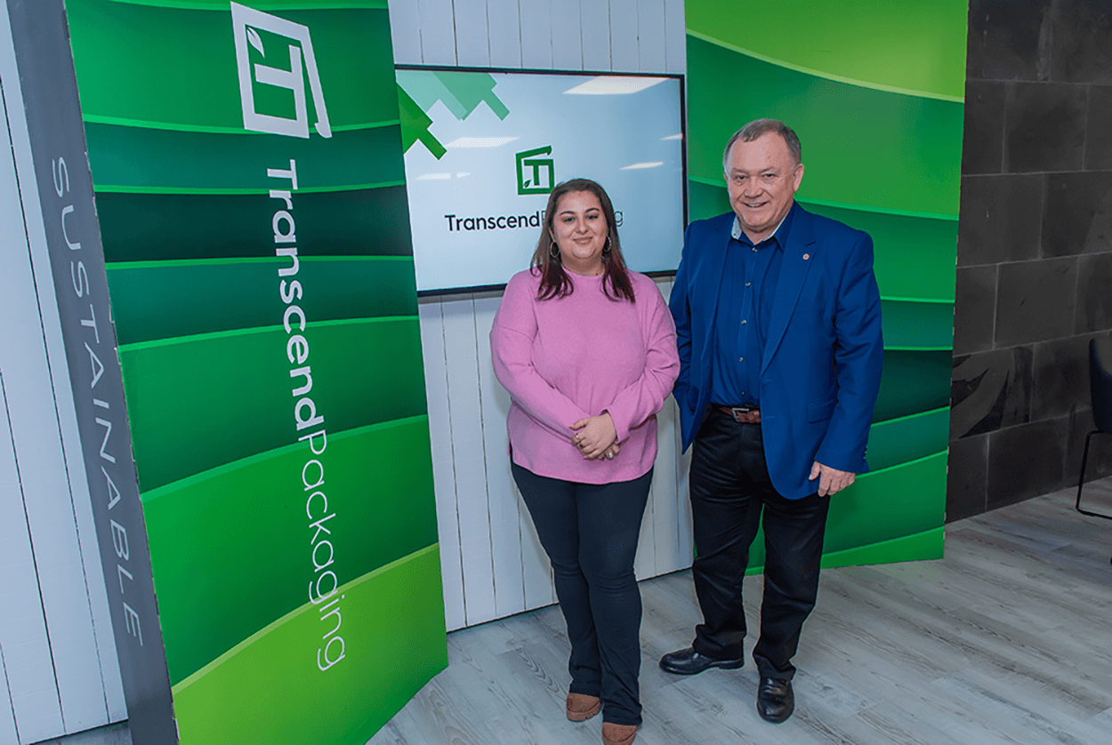 Two adults posing for a photo inside a green-and-blue branded office or event space, standing beside a large “Transcend Packaging” display.