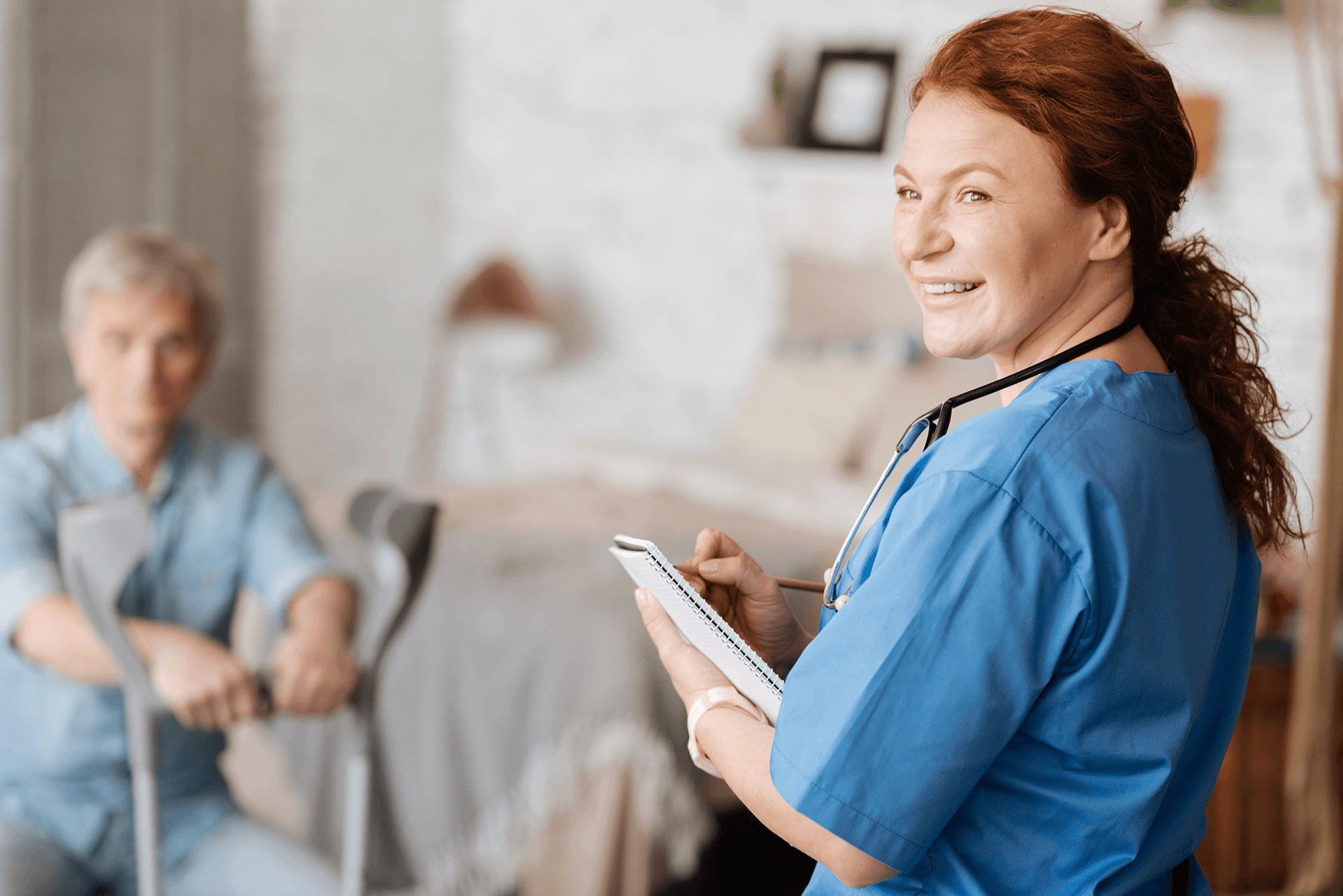 A healthcare professional in blue scrubs holding a notebook while supporting an older adult seated in a care setting.