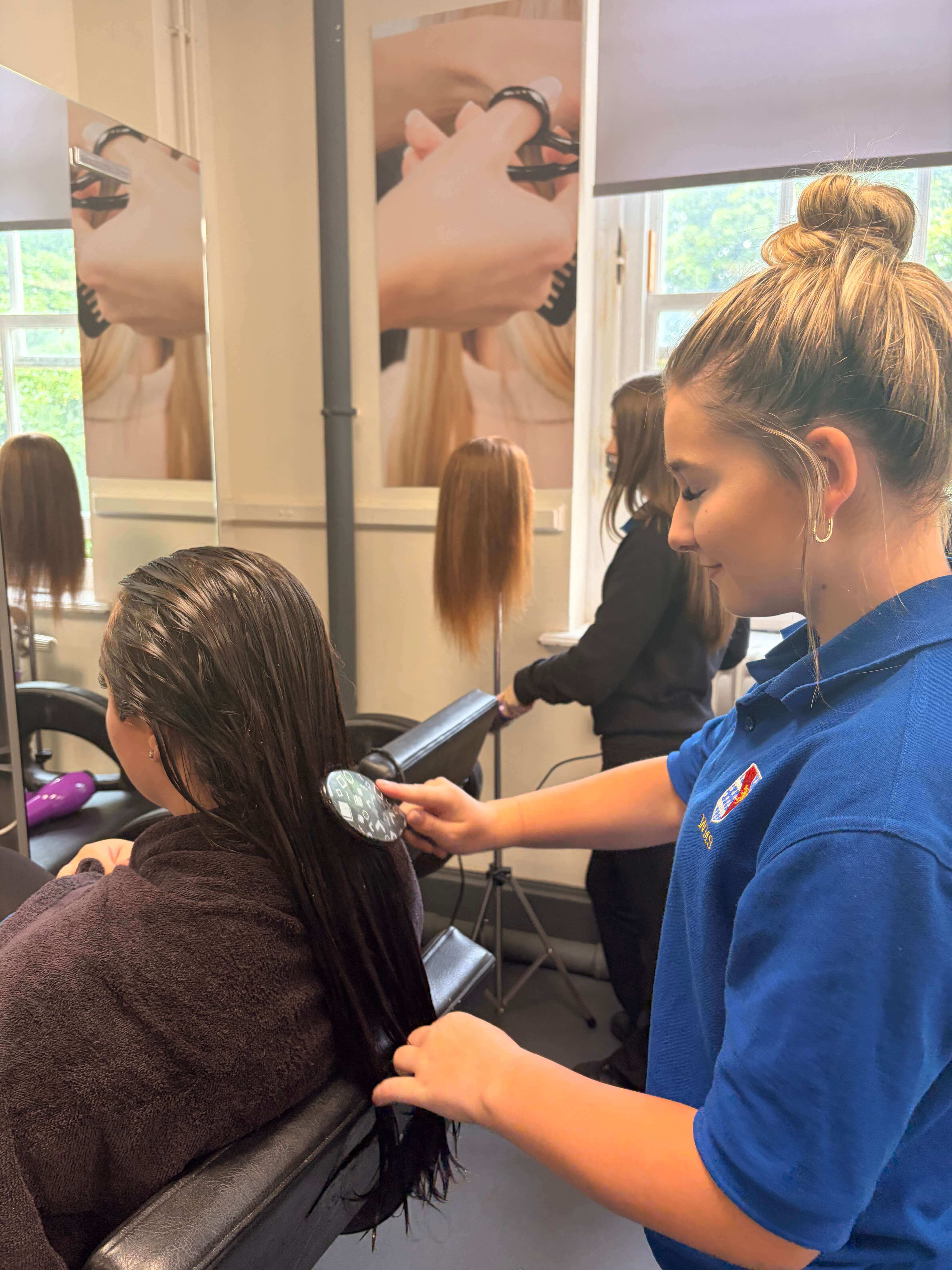 A hairdressing student in a blue polo shirt brushes a client's long, wet hair in a professional hair and beauty training salon.