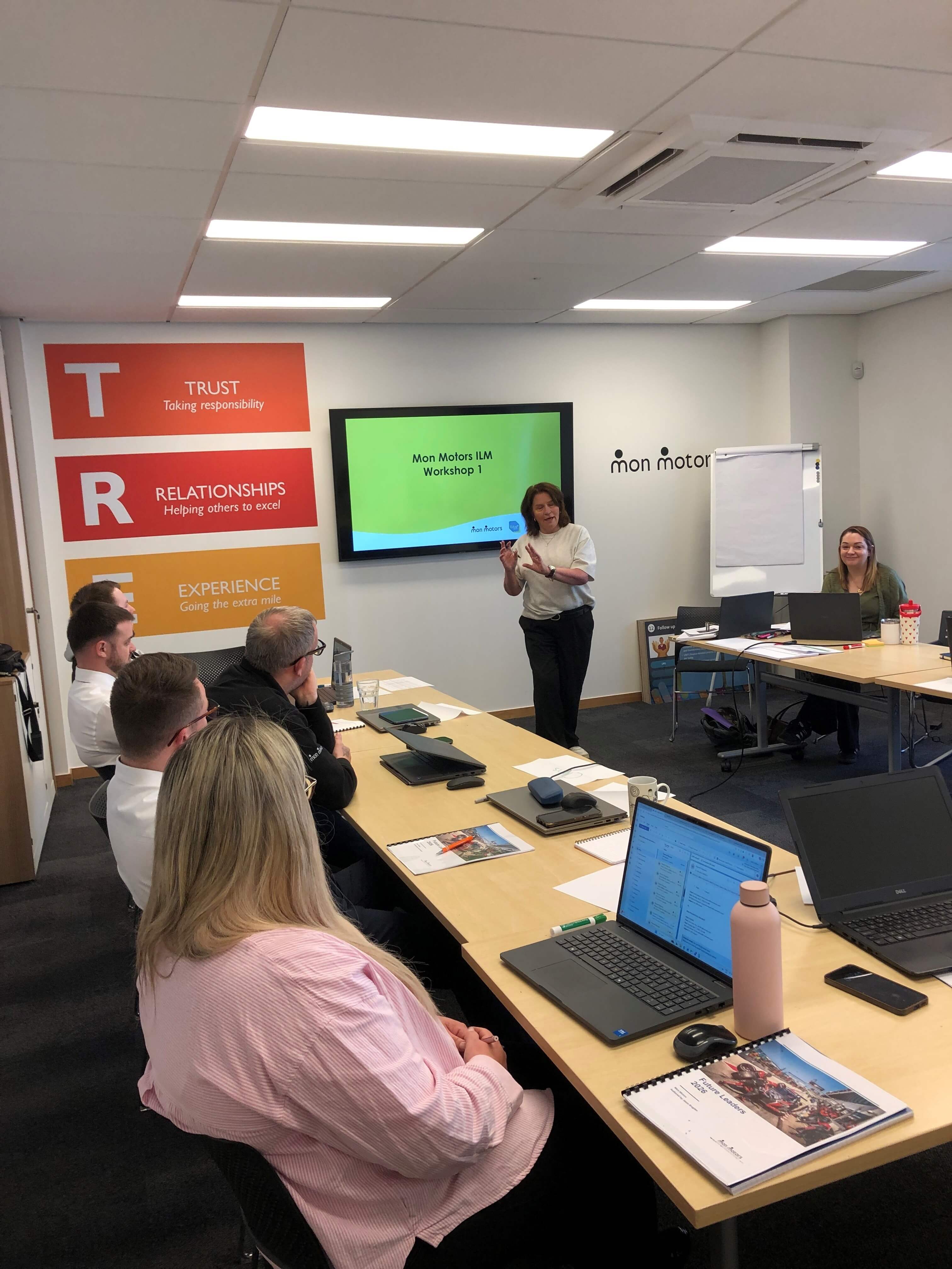 A trainer presenting a workshop to a group of colleagues seated around tables with laptops and notebooks in a meeting room.