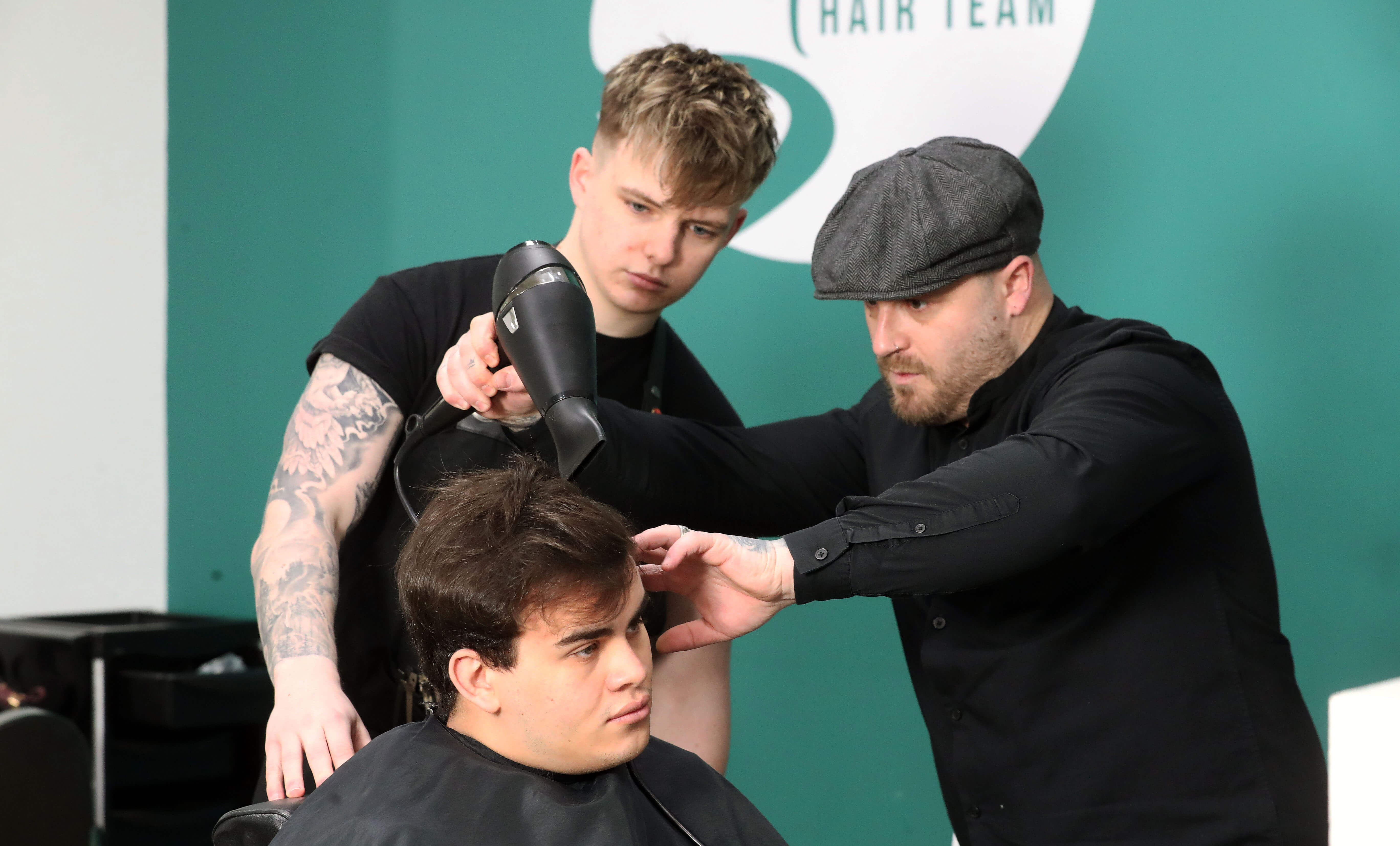A barber demonstrates hair-drying and styling techniques on a seated client while a trainee observes in a professional hair salon.