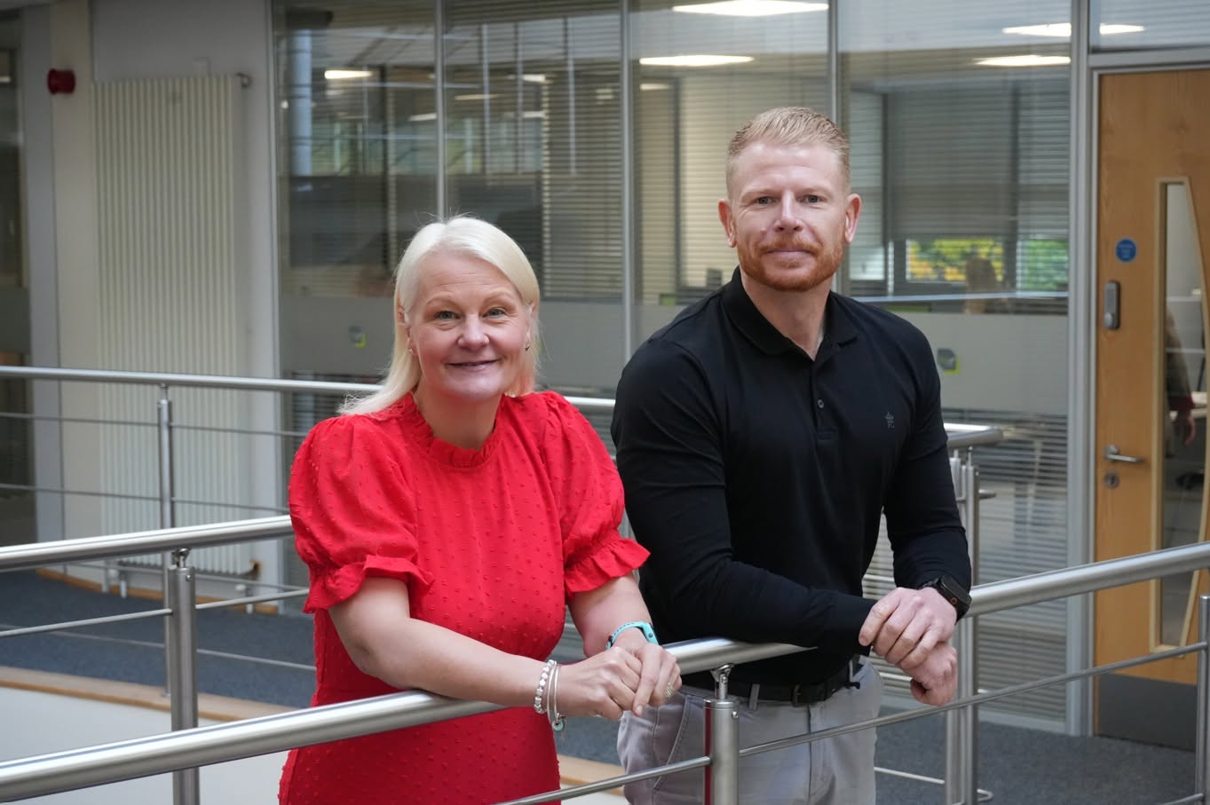 A woman in a bright red blouse and a man in a black polo shirt pose together, leaning against a metal railing in a modern office building.
