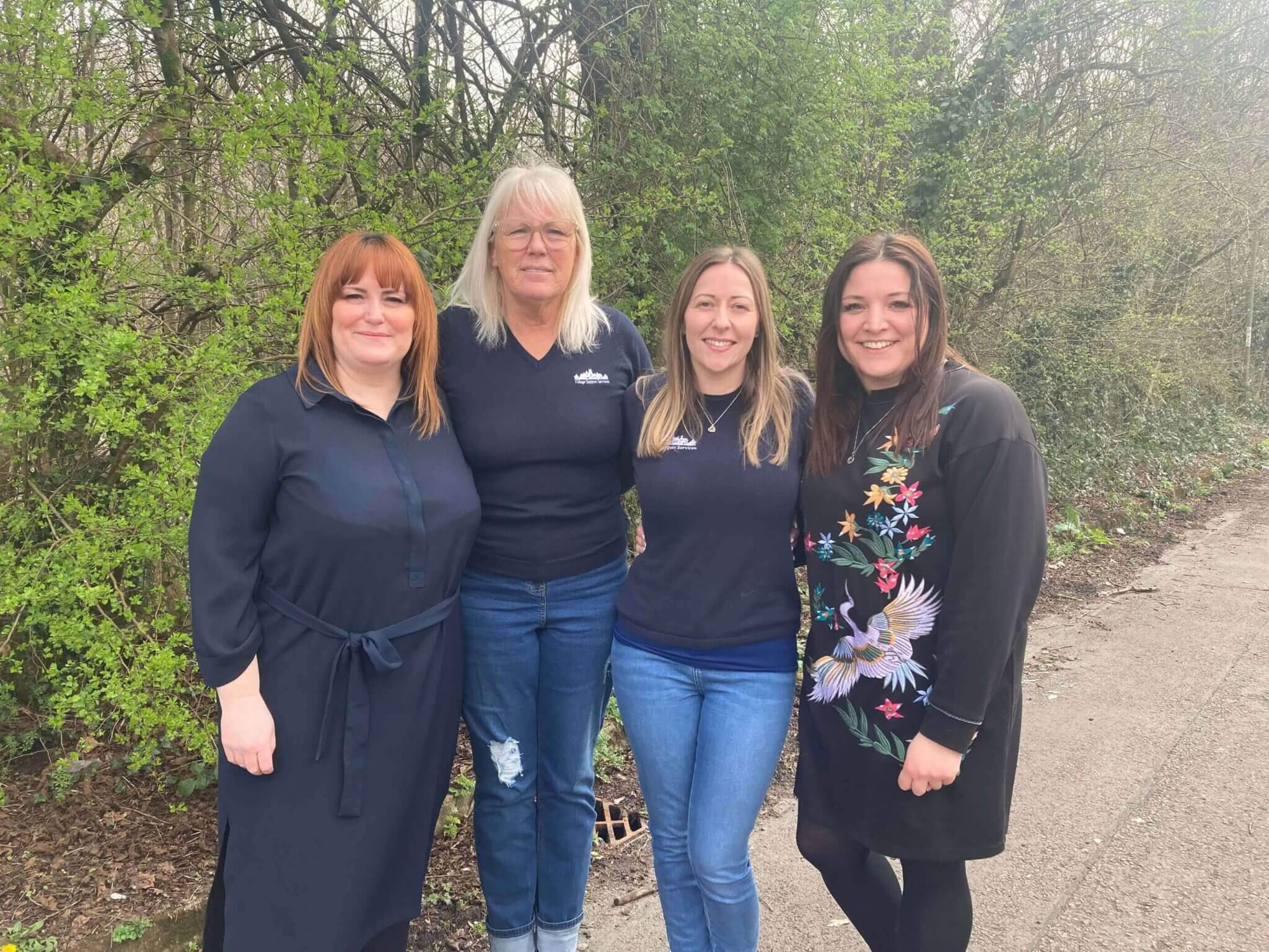 Four female colleagues standing together outdoors, smiling at the camera in a leafy setting.