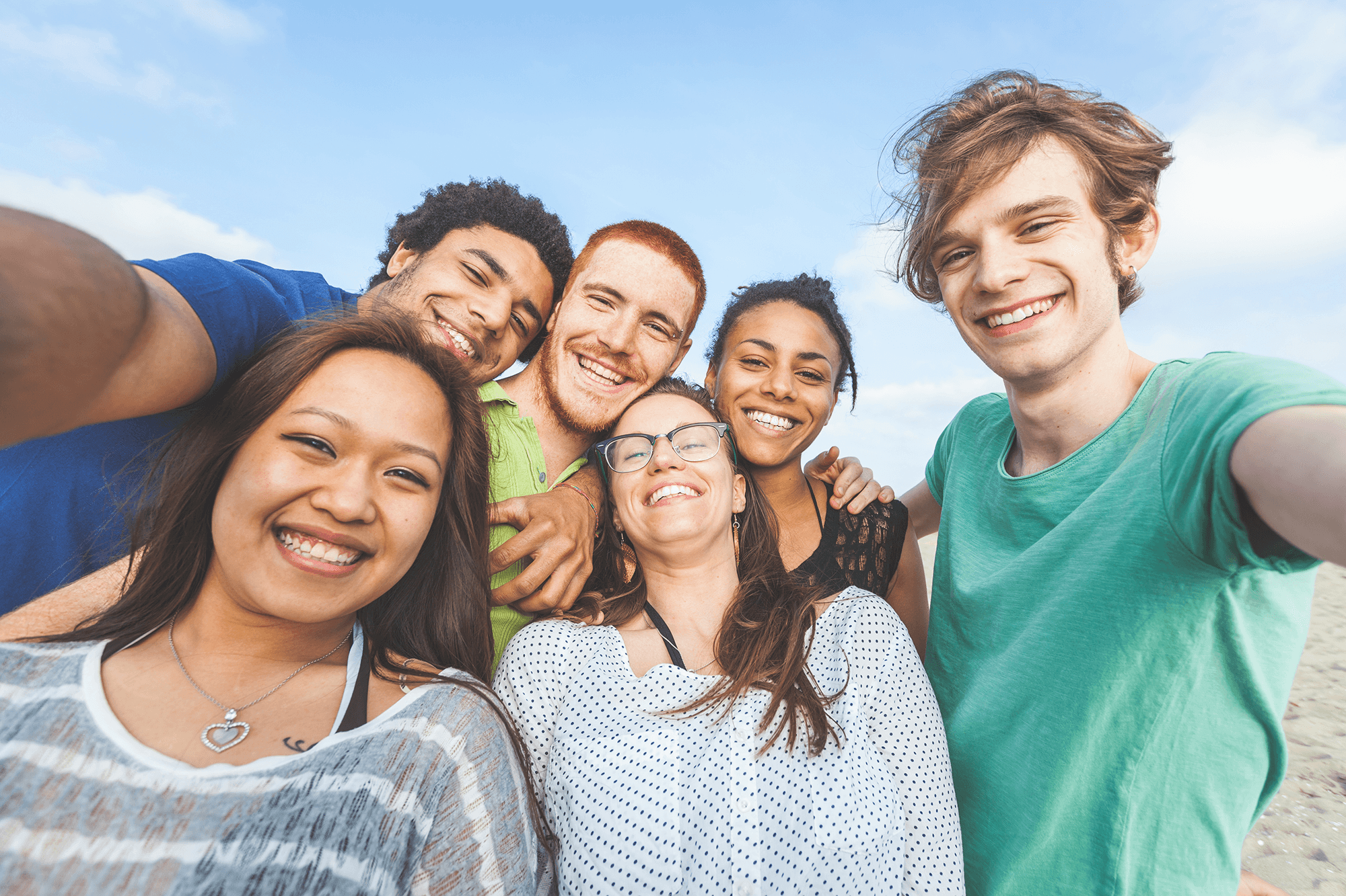 A diverse group of six young people smiling together for a group selfie outdoors on a sunny day.